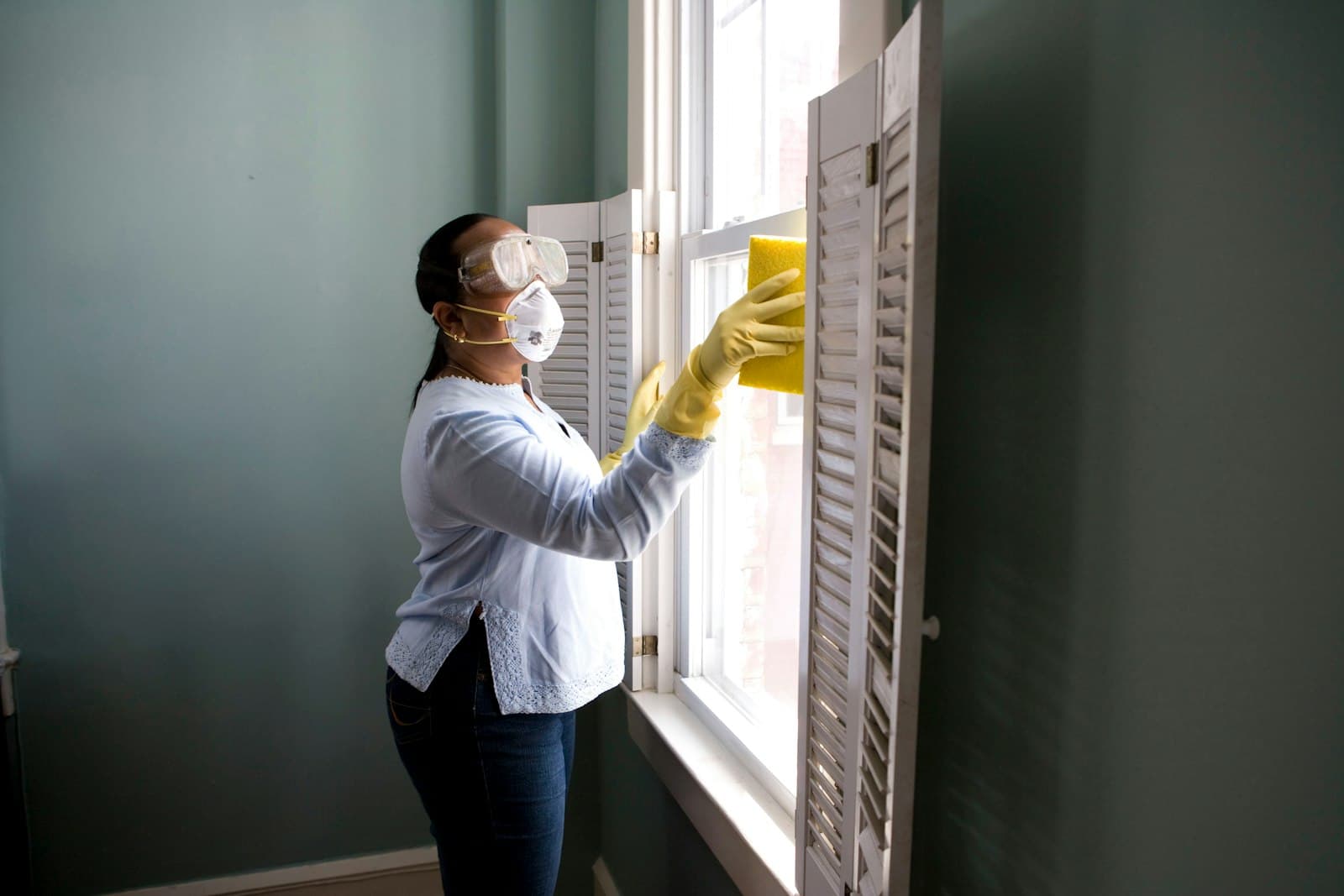 A professional cleaner wearing gloves carefully wiping white window shutters in a bright home